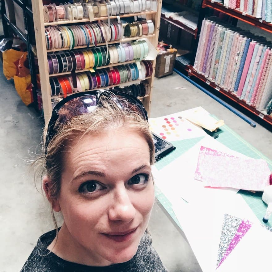 Kerri at the cutting table in her fabric warehouse, sunglasses pushed up on her head, with a rack of ribbon spools and shelves of fabric bolts behind her and half-cut bunting on the mat.