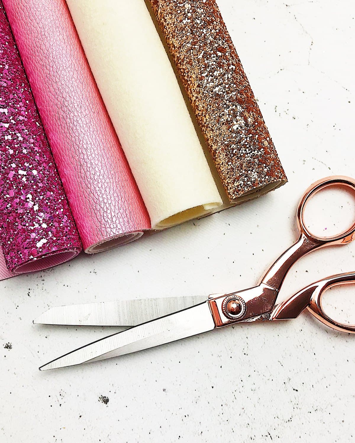 Rolls of glitter fabric and pink leather laid on a marble worktop next to a pair of rose gold dressmaking shears.